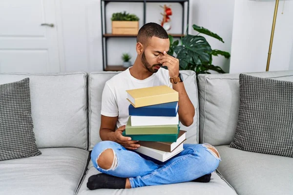 African american young man holding a pile of books sitting on the sofa tired rubbing nose and eyes feeling fatigue and headache. stress and frustration concept.