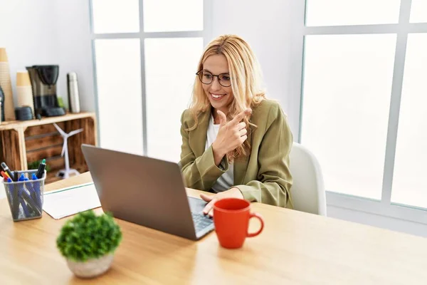 Beautiful blonde woman working at the office with laptop cheerful with a smile of face pointing with hand and finger up to the side with happy and natural expression on face