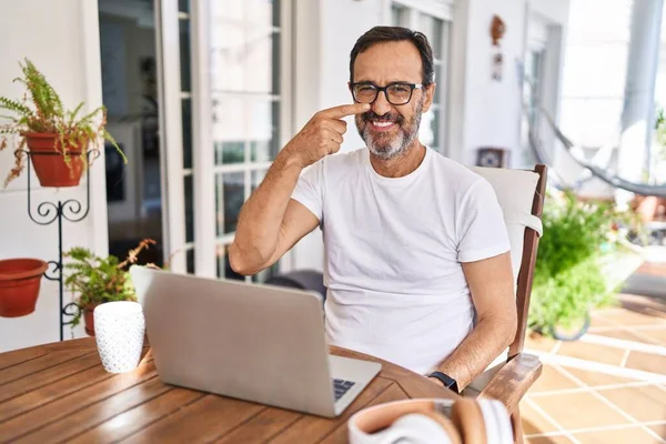Middle age man using computer laptop at home pointing with hand finger to face and nose, smiling cheerful. beauty concept