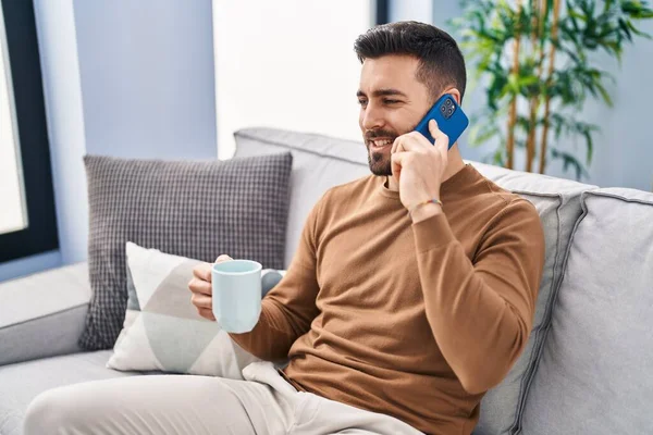 Young hispanic man drinking coffee and talking on the smartphone at home