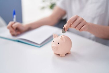 Young woman writing on notebook inserting coin on piggy bank at home