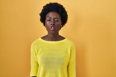African young woman standing over yellow studio making fish face with lips, crazy and comical gesture. funny expression. 