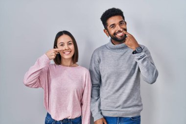 Young hispanic couple standing together pointing with hand finger to face and nose, smiling cheerful. beauty concept 