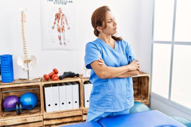 Middle age hispanic physiotherapist woman working at pain recovery clinic looking to the side with arms crossed convinced and confident 