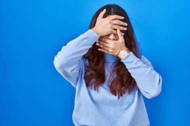 Hispanic young woman standing over blue background covering eyes and mouth with hands, surprised and shocked. hiding emotion 