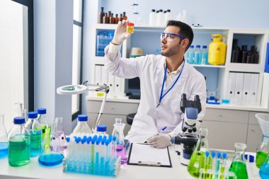 Young hispanic man scientist looking urine test tube writing report at laboratory