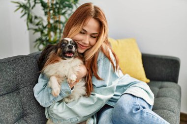 Genç beyaz kadın kanepede oturuyor ve evde köpeğe sarılıyor.