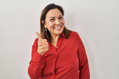 Hispanic mature woman standing over white background doing happy thumbs up gesture with hand. approving expression looking at the camera showing success. 