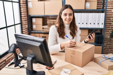 Genç ve güzel İspanyol kadın ecommerce iş işçisi. Ofisteki pakette akıllı telefon yazısını kullanıyor.