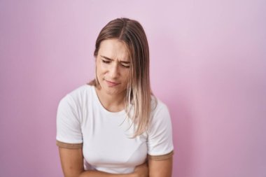 Blonde caucasian woman standing over pink background with hand on stomach because indigestion, painful illness feeling unwell. ache concept. 