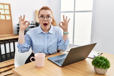 Ofiste bilgisayar laptopu kullanan genç kızıl saçlı kadın şaşırmış ve şok olmuş bir şekilde parmaklarıyla onay sembolü çiziyor. çılgın ifade 