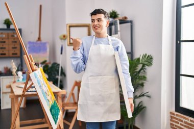 Non binary person at art studio pointing thumb up to the side smiling happy with open mouth 
