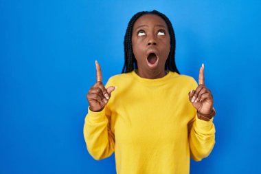 Beautiful black woman standing over blue background amazed and surprised looking up and pointing with fingers and raised arms. 
