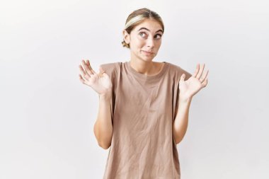 İzole edilmiş arka planda duran genç sarışın kadın elleri ve kolları havada, kafası karışmış bir ifade takınıyor. şüphe kavramı. 