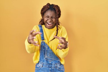 African woman standing over yellow background shouting frustrated with rage, hands trying to strangle, yelling mad 