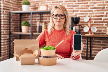 Sarışın kadın yemek yiyor. Akıllı telefon ekranı var. Düşünme tavrı ve kendine güvenen ayık yüz ifadesi var. 