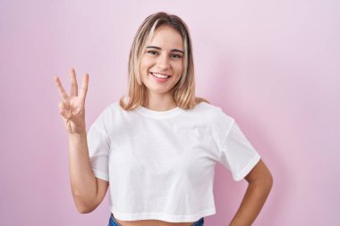 Young blonde woman standing over pink background showing and pointing up with fingers number three while smiling confident and happy. 