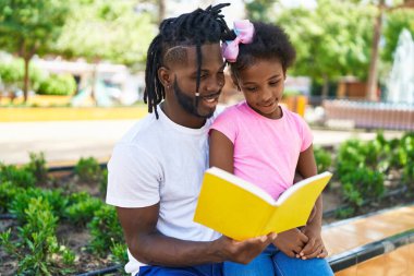 Father and daughter reading book sitting together on bench at park