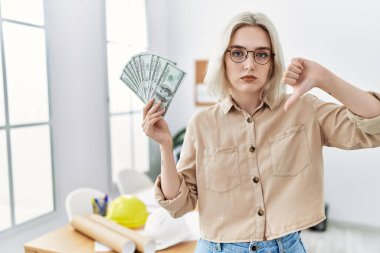 İnşaat ofisindeki genç, beyaz bir kadın elinde kızgın yüzle para tutuyor. Negatif işaretler baş parmakları aşağıda. Reddedilme kavramı. 