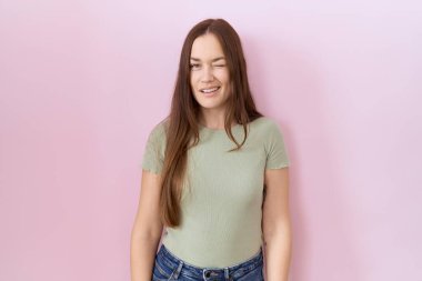 Güzel esmer kadın pembe arka planda duruyor. Seksi bir ifadeyle kameraya bakıyor. Neşeli ve mutlu bir yüzü var.. 