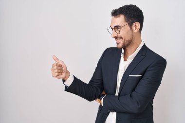 Handsome business hispanic man standing over white background looking proud, smiling doing thumbs up gesture to the side 