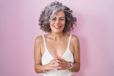 Middle age woman with grey hair standing over pink background hands together and fingers crossed smiling relaxed and cheerful. success and optimistic 