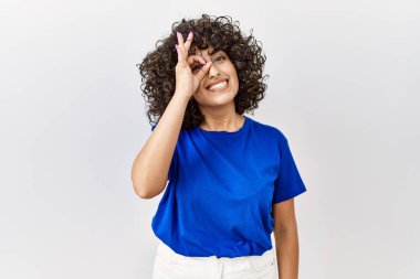 Ortadoğulu genç bir kadın izole arka planda duruyor. El hareketleriyle gülümsüyor, gözleriyle mutlu yüzle parmaklarının arasından bakıyor.. 