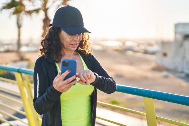 Middle age hispanic woman working out with smartphone at promenade