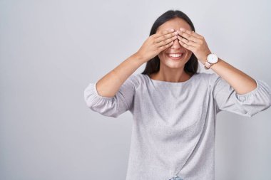 Young hispanic woman standing over white background covering eyes with hands smiling cheerful and funny. blind concept. 
