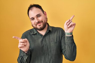 Plus size hispanic man with beard standing over yellow background smiling confident pointing with fingers to different directions. copy space for advertisement 