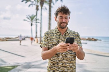 Genç İspanyol adam deniz kenarında akıllı telefon kullanarak gülümsüyor.