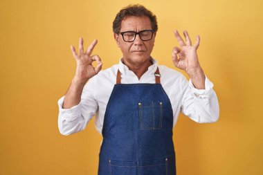 Middle age hispanic man wearing professional cook apron relax and smiling with eyes closed doing meditation gesture with fingers. yoga concept. 