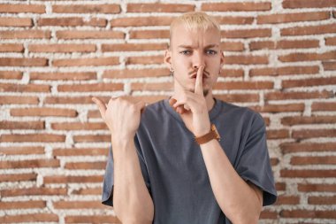 Kafkasyalı genç bir adam tuğla duvarın üzerinde duruyor ve parmakları yana doğru uzanmış bir şekilde sessiz olmak istiyor. sessizlik ve gizli konsept. 