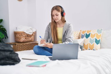 Young caucasian woman writing on notebook listening to music studying at bedroom