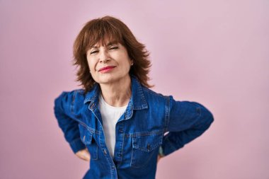 Orta yaşlı bir kadın pembe arka planda duruyor sırt ağrısı çekiyor, elleriyle dokunuyor, kas ağrısı çekiyor. 