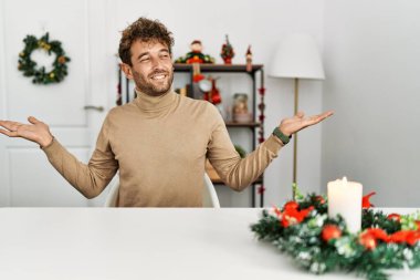 Noel süslemesi masasında oturan sakallı yakışıklı genç adam her iki elinde açık avuçlar, sunum ve reklam karşılaştırma ve denge gösteren gülümseme 