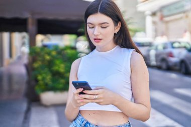 Beyaz bir kadın sokakta ciddi bir ifadeyle akıllı telefon kullanıyor.