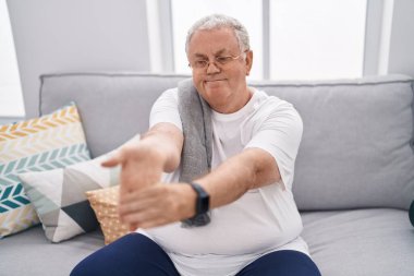 Orta yaşlı, kır saçlı, kollarını esneten adam evdeki kanepede oturuyor.