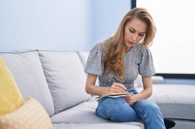 Evdeki kanepede oturan genç sarışın kadın deftere yazı yazıyor.