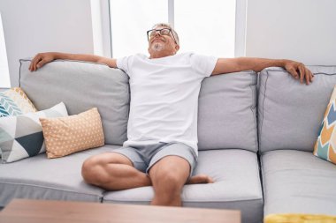 Orta yaşlı, gri saçlı, gülümseyen, evdeki kanepede rahatlamış bir adam.