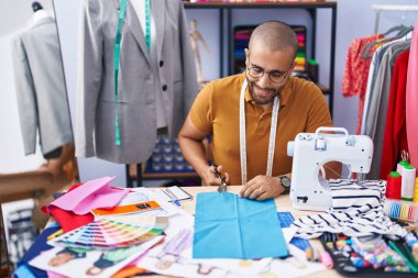 Young latin man tailor smiling confident cutting cloth at atelier