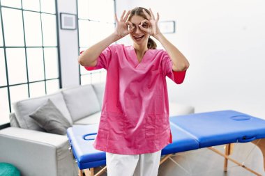 Genç sarışın kadın evde fizyoterapist olarak çalışıyor. Dürbün gibi jest yapıyor, gözleri parmaklarından bakıyor. çılgın ifade. 