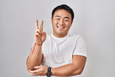 Young chinese man standing over white background smiling with happy face winking at the camera doing victory sign with fingers. number two. 