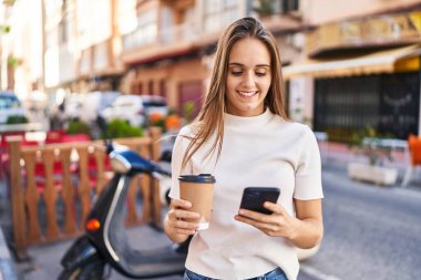 Young blonde woman using smartphone and drinking coffee at street