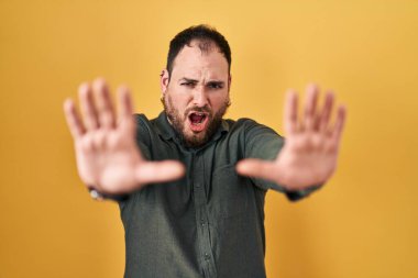 Plus size hispanic man with beard standing over yellow background doing stop gesture with hands palms, angry and frustration expression 