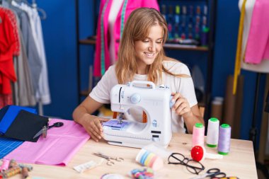 Young blonde girl tailor smiling confident using sewing machine at sewing studio