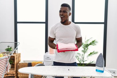 Young african man holding folded laundry after ironing angry and mad screaming frustrated and furious, shouting with anger. rage and aggressive concept. 