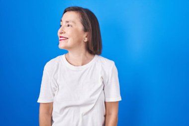 Orta yaşlı İspanyol bir kadın mavi arka planda duruyor. Yüzünde gülümseme, doğal bir ifade ile yan yana bakıyor. Kendinden emin gülen. 