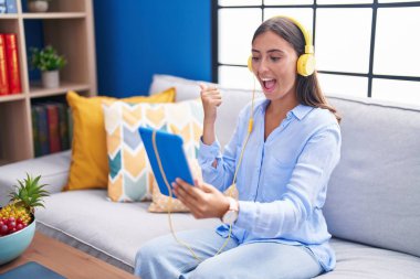 Young hispanic woman doing video call with tablet wearing headphones pointing thumb up to the side smiling happy with open mouth 