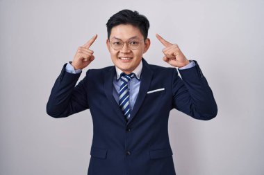 Young asian man wearing business suit and tie smiling pointing to head with both hands finger, great idea or thought, good memory 